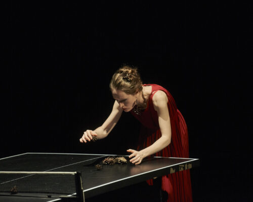 Eine Frau in einem roten Abendkleid beugt sich über eine Tischtennisplatte (Elena Rykova). Sie breitet Kegel darauf aus.