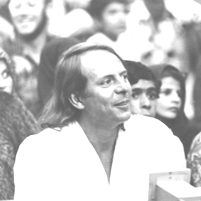 Auf dem Schwarzweißbild ist das Porträt eines Mannes zu sehen. Der Mann ist der Komponist Karlheinz Stockhausen. Er trägt ein weißes Hemd und schaut weg. Im Hintergrund sind weitere menschliche Figuren zu sehen.
