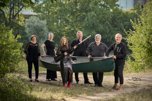 Vor der Kulisse der Natur stehen die sechs Sänger*innen der Neuen Vokalsolisten. Sie halten gemeinsam ein Boot. Die Frau im Vordergrund hält eine Schaufel.