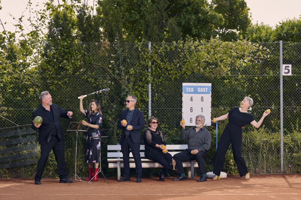 Auf dem Bild steht die Gruppe von sechs Sänger*innen der Neuen Vocalsolisten. Sie stehen auf dem Tennisplatz draußen und halten Bälle und Raketen in den Händen.