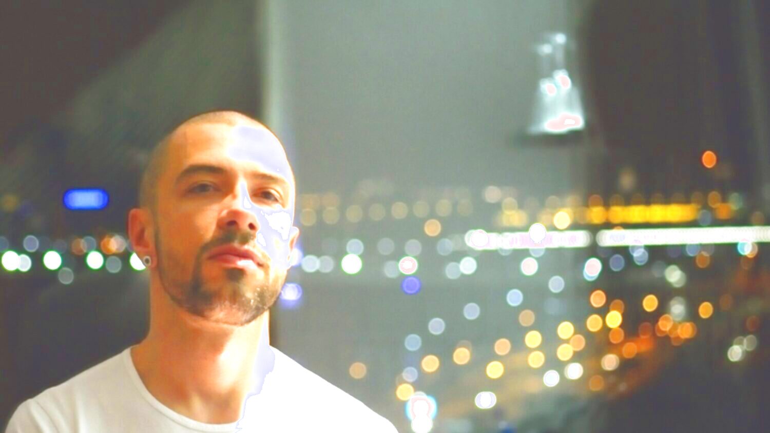 Das Porträt eines Mannes (Marko Nikodijević). Er blickt geradeaus und trägt ein weißes T-Shirt. Im Hintergrund sind die Lichter einer nächtlichen Stadt zu sehen.