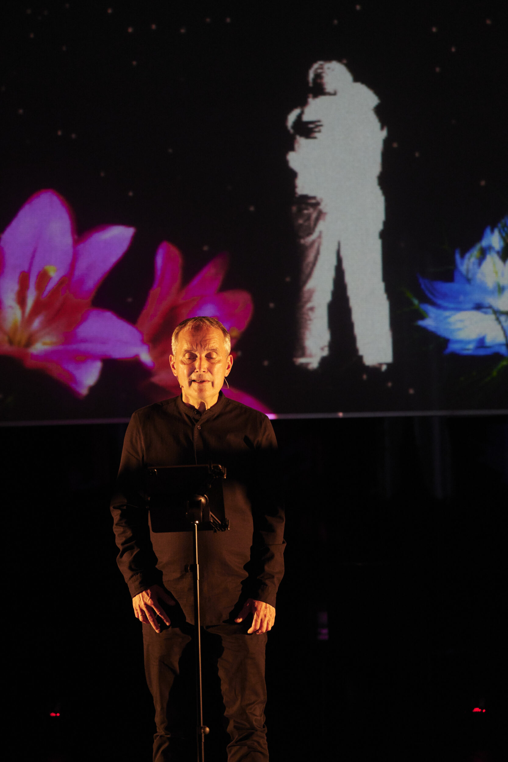 Auf der Bühne steht ein Sänger. Im Hintergrund ist eine Projektion mit zwei weiß gekleideten Personen zu sehen, die sich bunte Blumen umhängen.