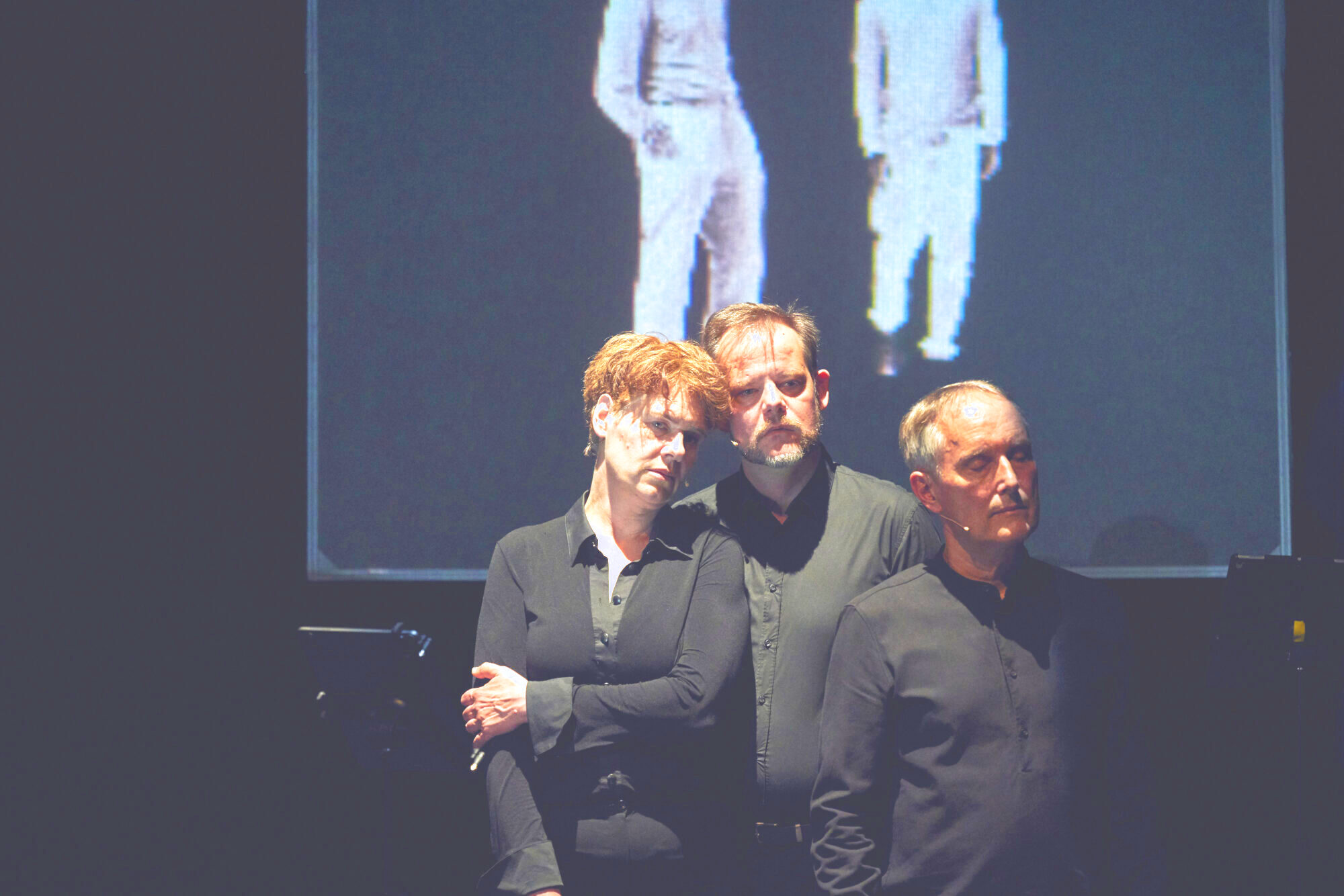 In der Mitte stehen drei Sänger*innen gegeneinander gelehnt. Im Hintergrund ist eine Projektion mit zwei weiß gekleideten Menschen zu sehen.