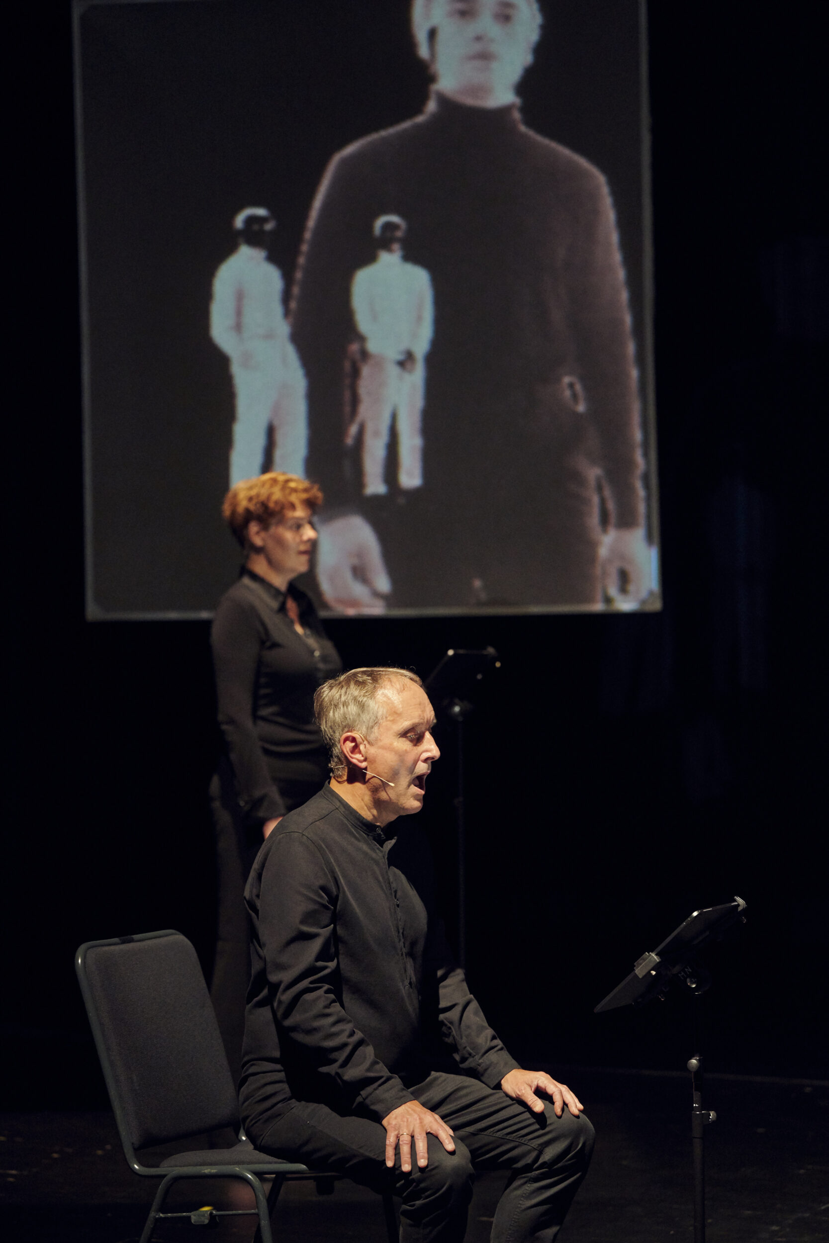 Eine Sängerin steht mit halber Drehung neben einem sitzenden Mann. Beide singen. Im Hintergrund ist eine Videoprojektion von drei Menschen zu sehen. Das Bild ist unscharf.