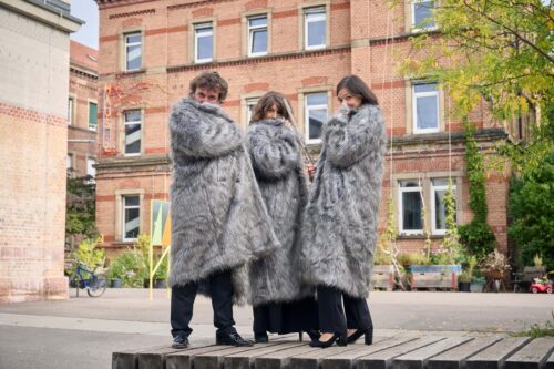 Das Bild zeigt zwei Frauen und einen Mann (TRIO vis-à-vis). Sie tragen Fellmäntel und verdecken damit ihre Gesichter. Im Hintergrund ist ein Gebäude zu erkennen.