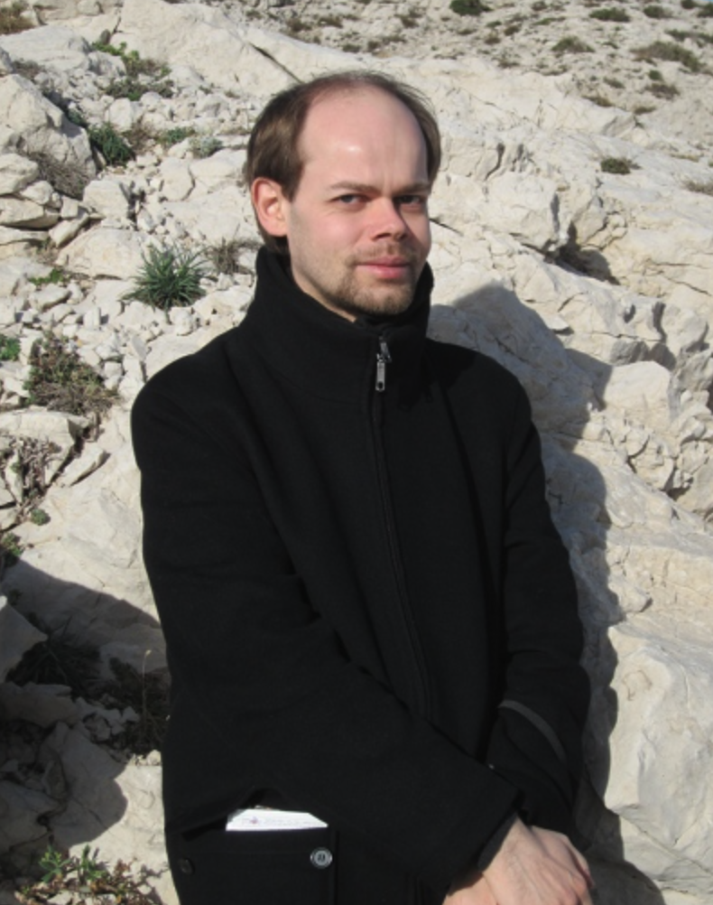 Porträt eines Mannes (Gilles Schuehmacher). Er sitzt im Sand, die Hände vor sich gefaltet. Er trägt ein schwarzes Oberteil.