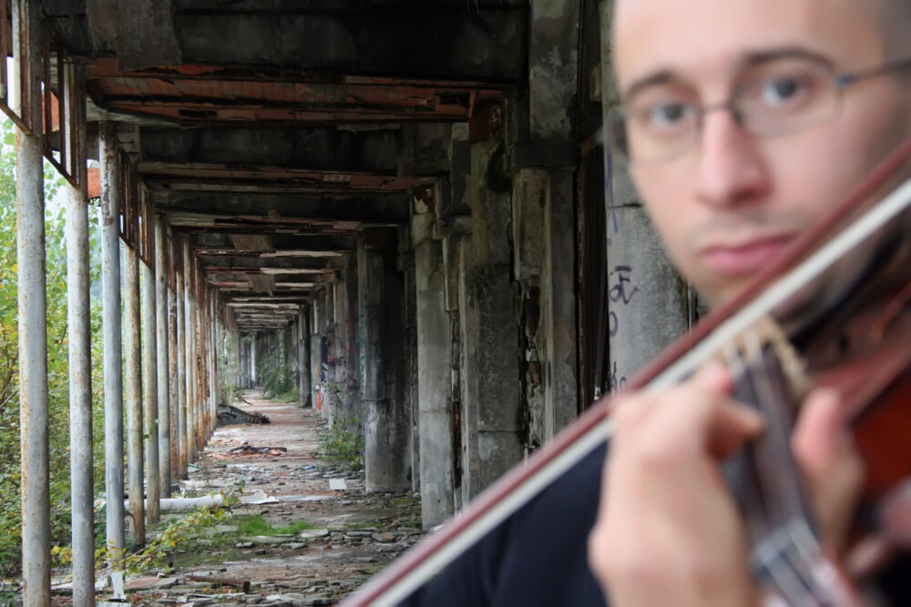 Zu sehen ist ein Portrait des violinist und violist Marco Fusi. Er ist sein Instrument spielend unscharf im Vordergrund zu sehen, im Hintergrund ist ein verfallener Durchgang zu sehen.